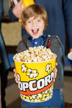 Portrait Of Excited Boy Offering Popcorn Bucket At Cinema