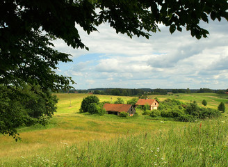 Obraz premium A rural spring view. Green meadows framed by branches in the foreground. In the background, an old red-brick farmhouse. Poland, Masuria.