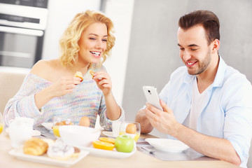 Happy couple eating breakfast and using smart phone
