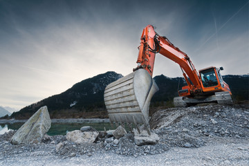 heavy organge excavator with shovel standing on hill with rocks - obrazy, fototapety, plakaty