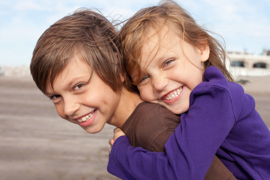 Happy elementary-age boy giving piggyback ride to laughing girl outdoors on a sunny summer day. Joyful childhood friendship, playful siblings bonding, and carefree fun 