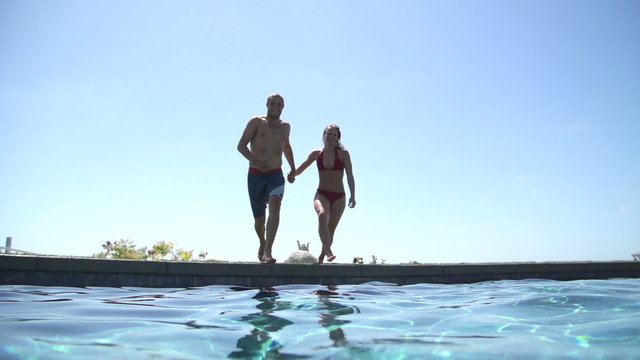 Young Happy Couple Holding Hands Running And Jumping Into Swimming Pool 