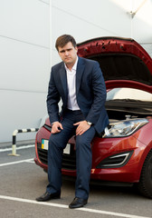 man in suit sitting on broken car with open hood