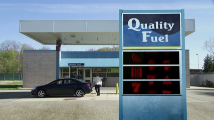 Woman gets out of car to pump gas.  Slot machine (fruit machine) icons spin on sign instead of prices.  They spin and stop on three sevens, showing prices are a gamble.