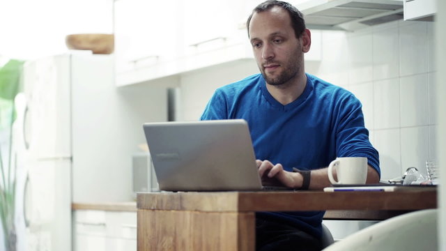 Young man using laptop and drinking coffee by table in kitchen 