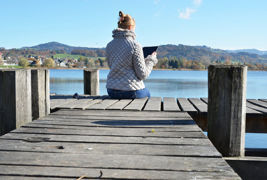Girl reading from a tablet on the wooden jetty against a lake. S
