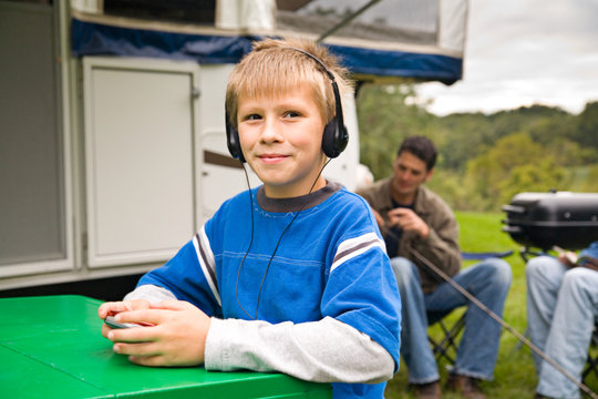 Camping: Boy Listens To Music While Dad Works With Fishing Pole