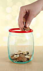 Female hand putting coins in glass bottle on bright background