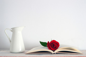 A vase beside an open book with a red rose on it.