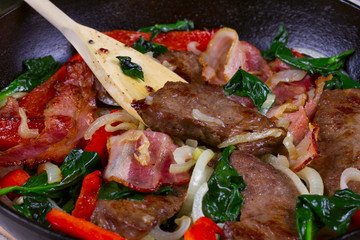 Frying pan with fried meat, bacon, red pepper and spinach