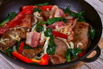 Frying pan with fried meat, bacon, red pepper and spinach
