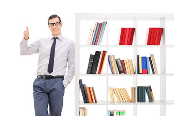 Young man giving thumb up and leaning on bookshelf