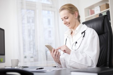 Female Doctor at her Table Browsing on her Phone - obrazy, fototapety, plakaty