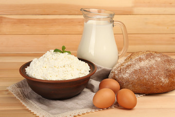 Cottage cheese in clay bowl with jug of milk,eggs and loaf of bread on wooden planks background