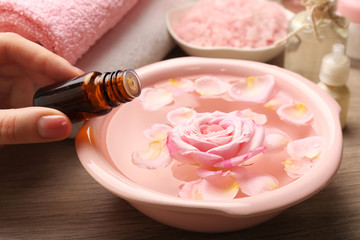Female hand with bottle of essence and bowl of spa water with flowers on wooden table, closeup