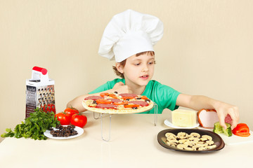 Little boy in chefs hat puts the ingredients on the pizza crust