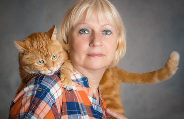 Beautiful aged woman hugging her red cat on grey background.