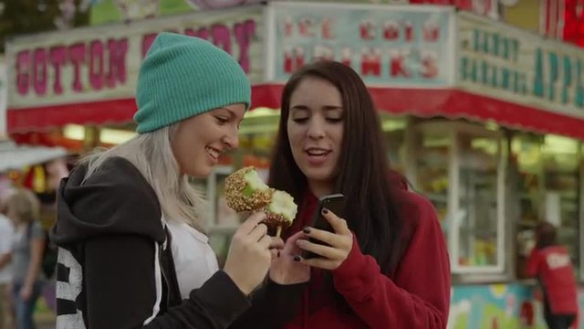 Medium shot of young women eating taffy apples and taking self-portraits with camera phone at amusement park / Salt Lake City, Utah, United States