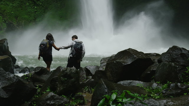 Slow motion wide panning shot of hiking couple admiring waterfall in rain forest / Arenal, La Fortuna, Costa Rica
