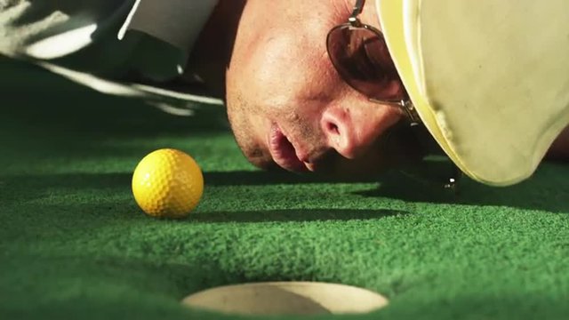CU Man lying on golf course green blowing golf ball towards hole, Orem, Utah, USA