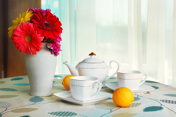 Colorful gerbera in vase with teapot, cups and lemons on table on curtains background