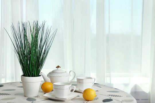 Green plant with teapot, cups and lemons on table on curtains background