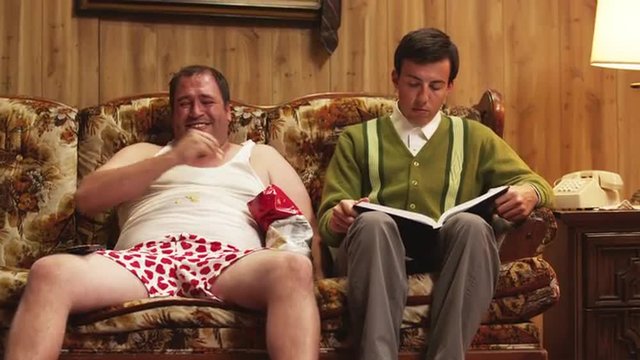 MS Obese man wearing underwear sitting on sofa next to geek reading book Orem, Utah, USA