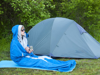 A man sits in a sleeping bag near the tent.
