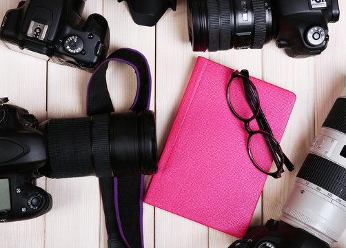 Modern cameras with glasses and pink diary on wooden table, top view