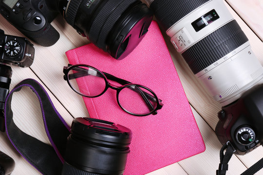 Modern cameras with glasses and pink diary on wooden table, closeup