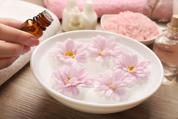 Female hand with bottle of essence and bowl of spa water with flowers on wooden table, closeup