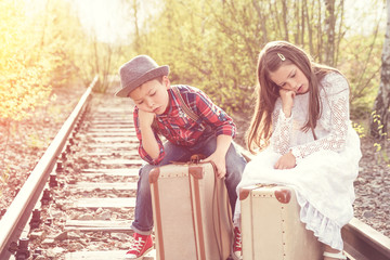 Fototapeta na wymiar Mädchen und Junge warten auf Koffern sitzend an alten Bahngleis