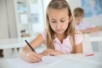 Cute schoolgirl writing on notebook, in classroom