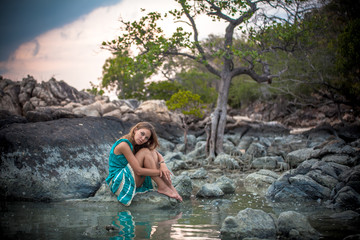 Fototapeta premium Young beautiful woman in long turquoise dress sitting on a stone