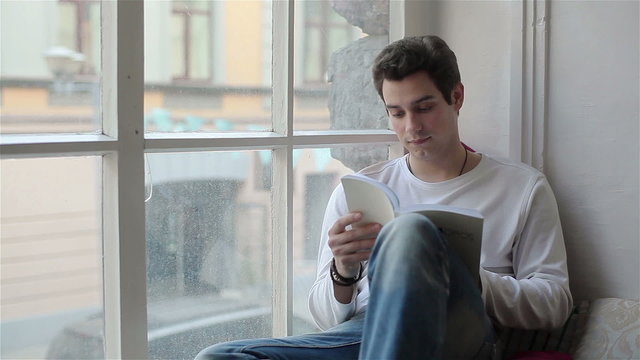Young man reading a book sitting by the window