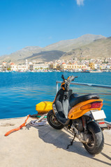 Fototapeta premium Orange scooter standing on wharf of Greek harbour with city budi