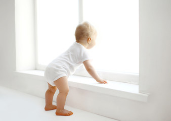 Cute baby at home in white room stands near window