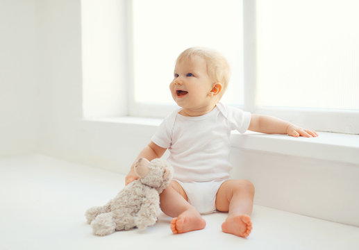 Cute smiling baby with teddy bear toy sitting at home in white r