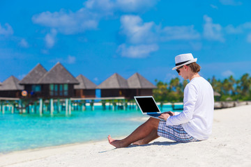 Young man with laptop at tropical beach near water villa
