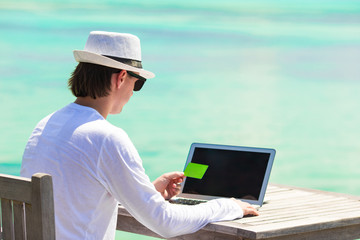 Young man working on laptop with credit card at tropical beach