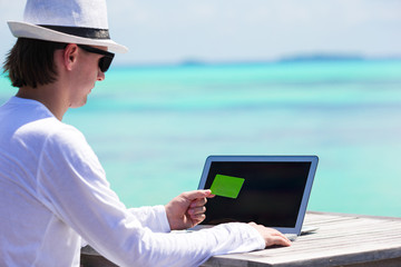 Young man working on laptop with credit card at tropical beach