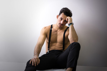 Shirtless athletic young man with suspenders sitting on floor