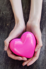 Obraz premium Beautiful female hands holding pink plastic heart over old woode