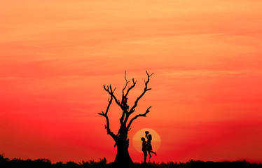 Silhouette of couple in love with big sunset and dead tree.