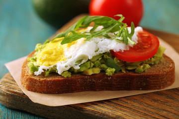 Tasty sandwich with egg, avocado and vegetables on cutting board, on color wooden background