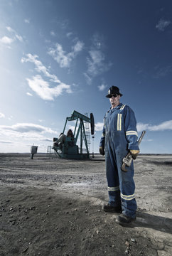 A man in overalls and hard hat at a pump jack in open ground at an oil extraction site.