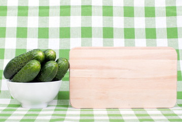 Cucumbers in a white bowl on a checkered napkin beside cutting b