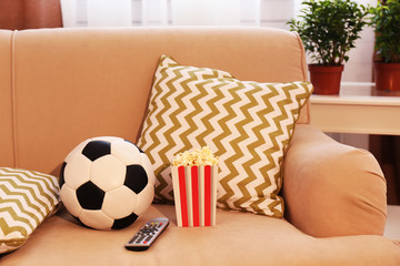Soccer ball, remote control and box of popcorn on comfortable sofa, indoors