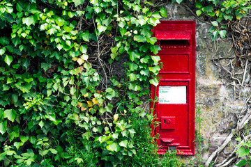 Red Post Box Mounted on a old Crumbling Wall. Overgrown Plants