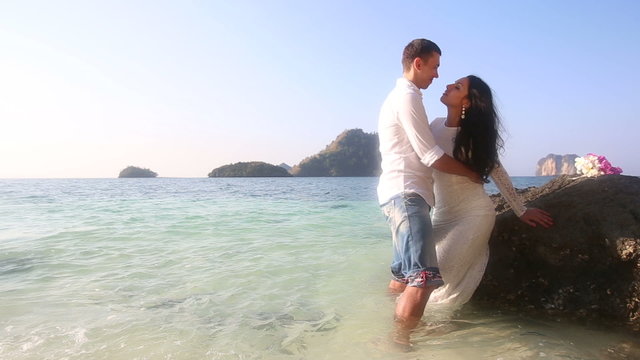 bride and groom stand in transparent sea leaning on rock